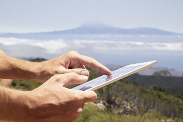 Spanien, Kanarische Inseln, La Gomera, Hände eines Wanderers, der ein digitales Tablet benutzt, mit dem Vulkan Teide im fernen Hintergrund - MAMF00895