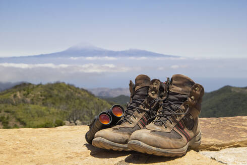 Spanien, Kanarische Inseln, La Gomera, Fernglas und ein Paar braune Wanderschuhe liegen auf dem Gipfel des Garajonay mit dem Vulkan Teide im Hintergrund - MAMF00898