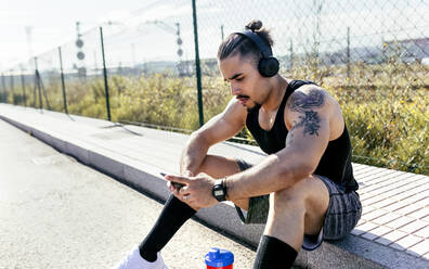 Sporty young man with smartphone and headphones sitting at the roadside - MGOF04132
