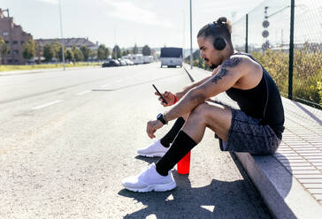 Sportlicher junger Mann mit Smartphone und Kopfhörern am Straßenrand sitzend - MGOF04133