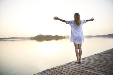 Rückansicht einer reifen Frau, die auf einem Steg an einem See bei Sonnenaufgang steht - PNEF02174