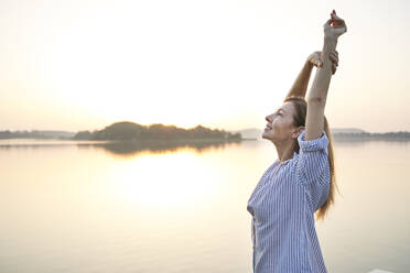 Happy mature woman stretching at a lake at sunrise - PNEF02175