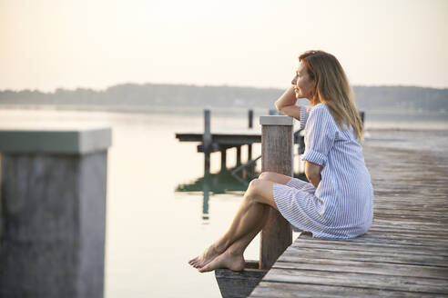 Ältere Frau sitzt auf einem Steg an einem See bei Sonnenaufgang - PNEF02181