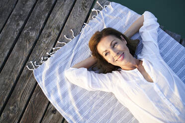 Lächelnde reife Frau auf einem Handtuch liegend auf einem Steg an einem See - PNEF02192