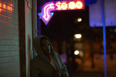 African-American man wearing hoodie under neon sign at night - CAVF65512