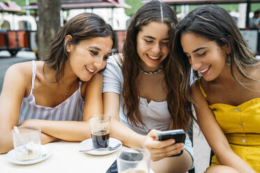 Three happy female friends sharing cell phone at a cafe in the city - MPPF00172