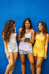 Drei glückliche Freundinnen stehen vor einer blauen Wand und teilen sich ein Tablet - MPPF00178