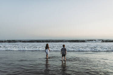 Siblings walking into the ocean staring at the horizon at sunset - CAVF65524