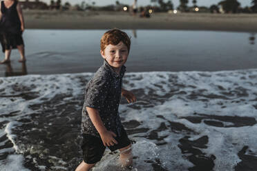 Young boy looking at camera while walking into the ocean at sunset - CAVF65525