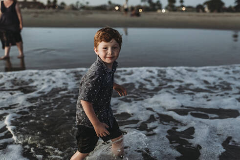 Junge schaut in die Kamera, während er bei Sonnenuntergang ins Meer geht - CAVF65525