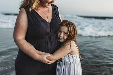 Seitenansicht einer glücklichen jungen Tochter, die ihre Mutter in der Abenddämmerung im Meer umarmt - CAVF65529