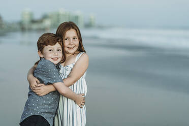 Porträt von Bruder und Schwester, die sich in der Abenddämmerung am Strand umarmen - CAVF65532