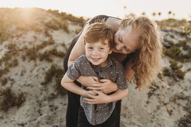 Vorderansicht der Mutter, die ihren rothaarigen Sohn im Kindergartenalter bei Sonnenuntergang umarmt - CAVF65537