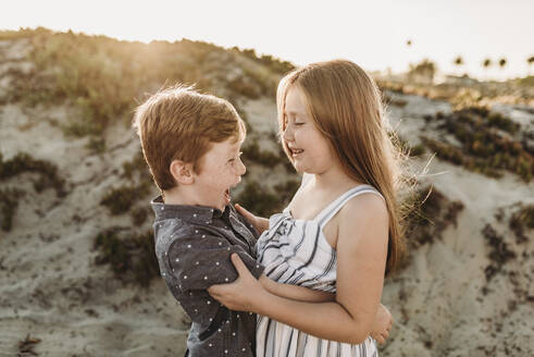 Seitenansicht eines jungen Bruders und einer jungen Schwester, die sich am Strand umarmen und lachen - CAVF65546