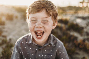 Vorderansicht eines kleinen Jungen im Kindergartenalter, der bei Sonnenuntergang am Strand lacht - CAVF65548