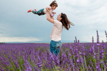 Frau wirft kleines Mädchen in die Luft zwischen Lavendelfeldern im Sommer - CAVF65569