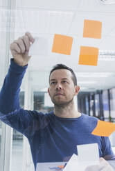 Mature businessman writing on sticky notes at glass pane in office - CAVF65591