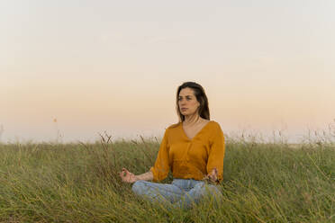 Junge Frau sitzt auf einer Wiese und meditiert am Abend - AFVF04068