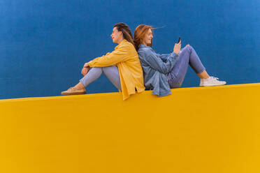 Junge, schöne, sorglose Frauen, die an einer orangefarbenen Wand sitzen, rothaarige Frau, die ein Smartphone benutzt - AFVF04075