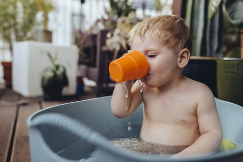 Kleiner Junge sitzt in der Babywanne auf dem Balkon und trinkt Wasser - MFF04898