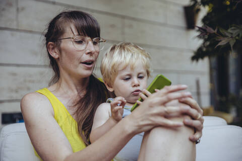 Kleiner Junge sitzt auf dem Schoß der Mutter und schaut auf sein Smartphone, lizenzfreies Stockfoto