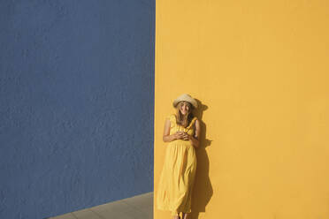 Frau in einem gelben Kleid mit Smartphone vor gelben und blauen Wänden - AHSF00962