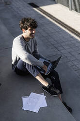 Lächelnder Mann sitzt auf einer Außentreppe mit Papieren und Laptop - GIOF07243