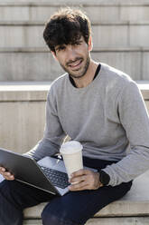 Mann sitzt auf einer Außentreppe mit Kaffee zum Mitnehmen und Laptop - GIOF07246