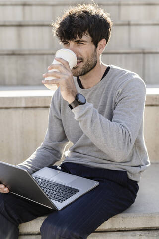 Mann sitzt auf einer Außentreppe mit Kaffee zum Mitnehmen und Laptop, lizenzfreies Stockfoto