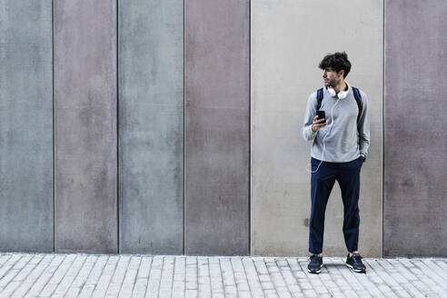 Mann mit Mobiltelefon und Kopfhörern vor einer Wand stehend - GIOF07265