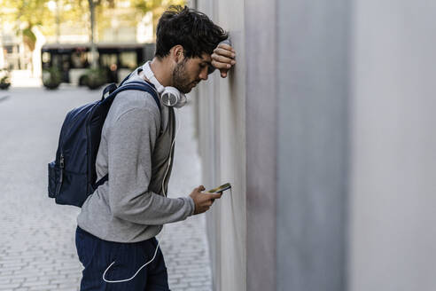 Mann lehnt an einer Wand und überprüft sein Mobiltelefon - GIOF07270