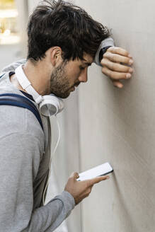Mann lehnt an einer Wand und überprüft sein Mobiltelefon - GIOF07271