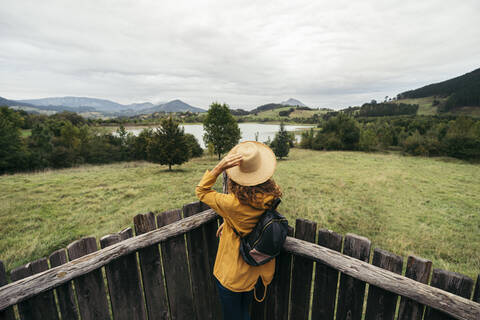 Junge Frau mit gelbem Mantel und Rucksack, die einen Hut in der Hand hält und auf einem Holzbalkon die Seenlandschaft betrachtet, lizenzfreies Stockfoto