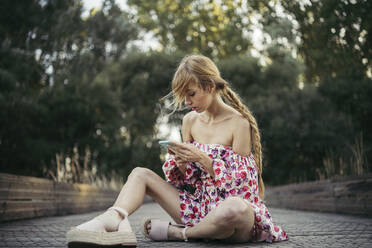 Junge Frau im Sommerkleid mit Blumenmuster sitzt auf der Promenade und schaut auf ihr Handy - MTBF00021