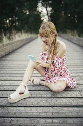 Porträt einer überraschten jungen Frau, die an der Strandpromenade sitzt und auf ihr Smartphone schaut - MTBF00024