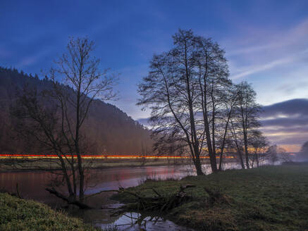 Germany, Bavaria, Silhouettes of trees against vehicle light trails stretching along shore of Naab river at dusk - HUSF00085