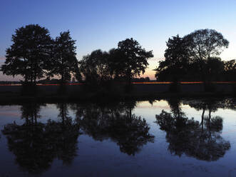 Deutschland, Bayern, Silhouetten von Bäumen, die sich in einem glänzenden See in der Abenddämmerung spiegeln - HUSF00101