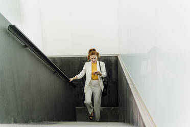 Businesswoman in white pant suit, ascending stairs, using smartphone - ERRF01800