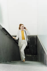 Businesswoman in white pant suit, ascending stairs, using smartphone - ERRF01801