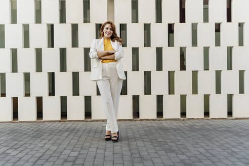 Geschäftsfrau in weißem Hosenanzug, vor einer Fassade stehend, mit verschränkten Armen - ERRF01815