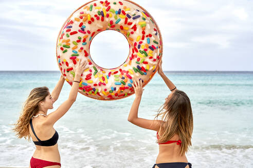 Two young women standing in front of the sea holding aloft a big inflatable ring - EPF00604