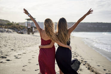 Two young women standing arm in arm on a beach with arms outstreched - EPF00627