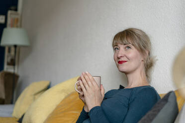 Porträt einer lächelnden Frau, die mit einer Tasse Kaffee auf der Couch sitzt - KNSF06823