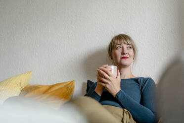 Portrait of woman sitting on the couch with cup of coffee looking at distance - KNSF06824
