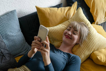 Porträt einer lächelnden Frau, die auf der Couch liegt und ein Smartphone benutzt - KNSF06826