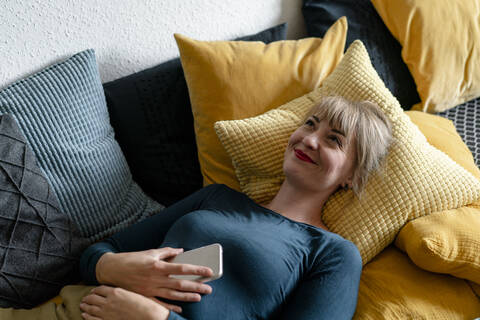 Porträt einer glücklichen Frau auf der Couch liegend mit Smartphone, lizenzfreies Stockfoto