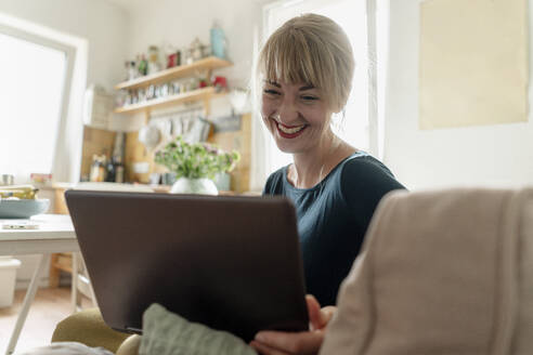 Portrait of happy woman sitting in the kitchen using laptop - KNSF06842