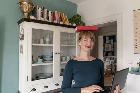 Portrait of smiling woman standing with laptop in the kitchen balancing book on her head - KNSF06843