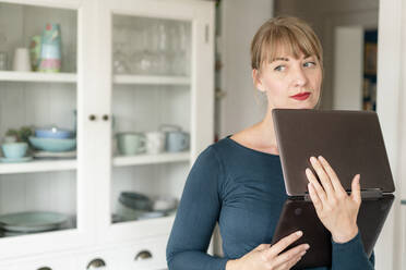 Porträt einer nachdenklichen Frau, die mit einem Laptop in der Küche steht und in die Ferne schaut - KNSF06846