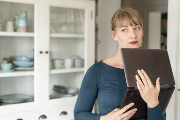 Portrait of pensive woman standing with laptop in the kitchen looking at distance - KNSF06846